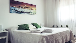 Un lit bien fait, recouvert d’une courtepointe blanche, avec deux oreillers et une pile de serviettes pliées. Un tableau représentant une gondole sur l’eau est accroché au mur, au-dessus du lit.