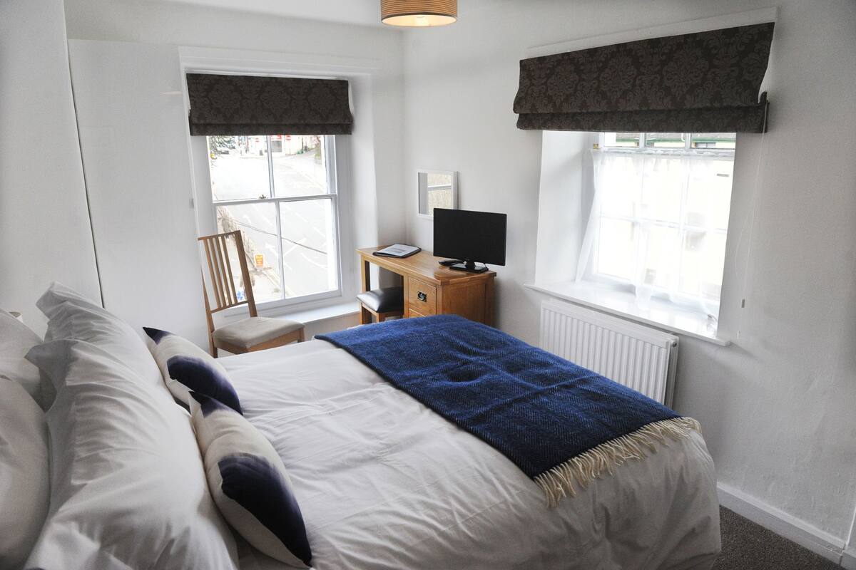 Ein Schlafzimmer mit einem Bett, einem Schreibtisch mit Computer und zwei mit Vorhängen versehenen Fenstern.