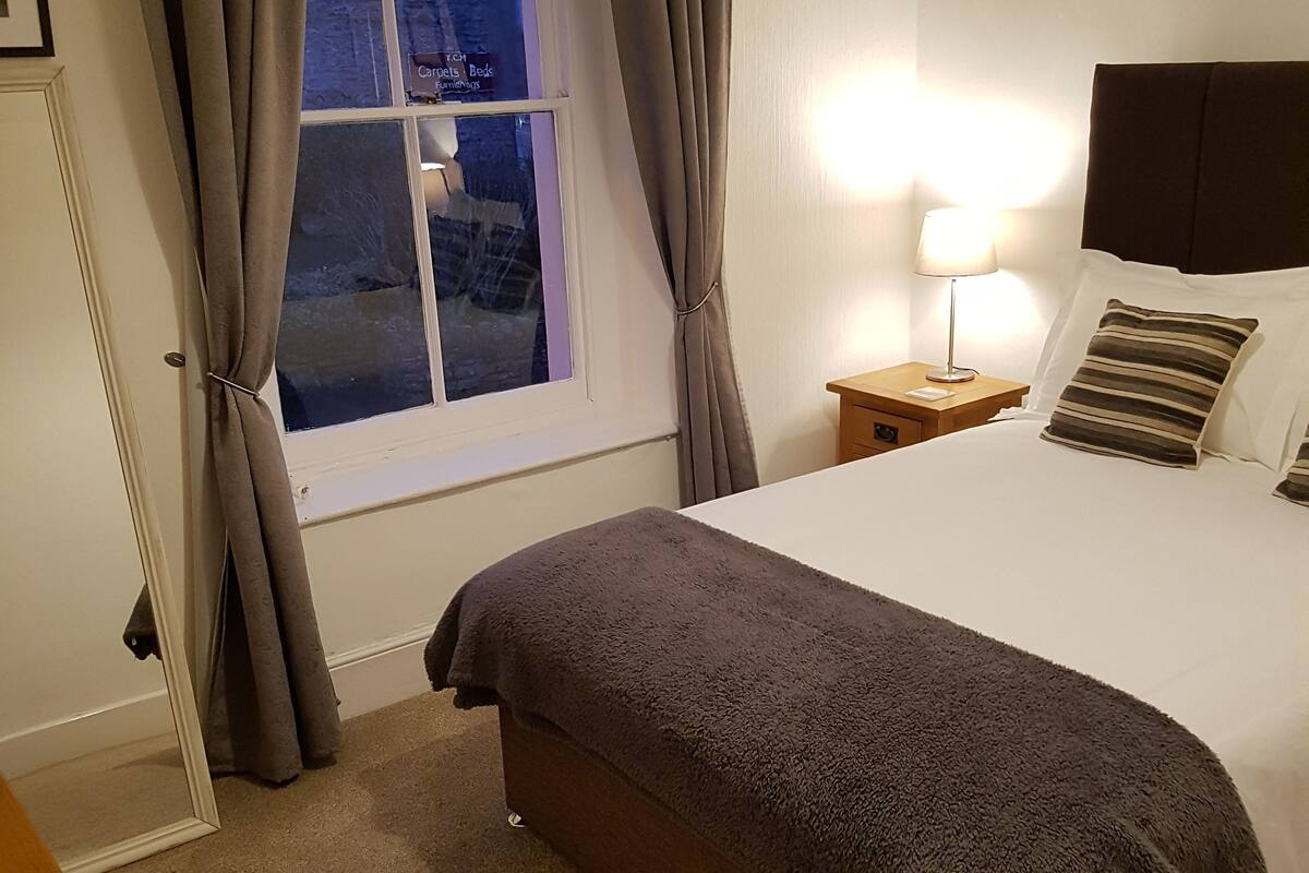 Una cama bien tendida con un edredón oscuro, una lámpara de noche y una ventana con cortinas.