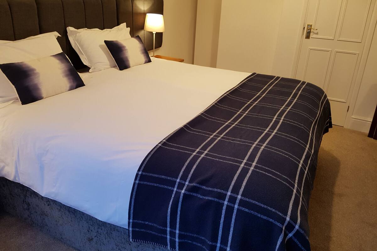 Una cama bien hecha con una colcha escocesa, un cabecero y una lámpara de noche.