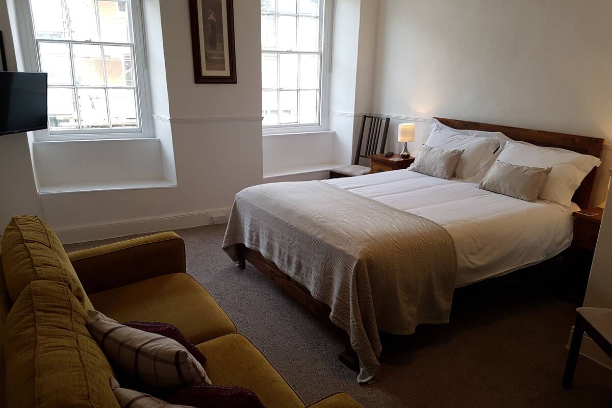 Un dormitorio con cama, sofá amarillo, mesita de noche y ventana.