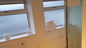 Baño con inodoro, lavabo y ducha. Cuenta con ventanas con persianas, un perchero y un pequeño estante con toallas.