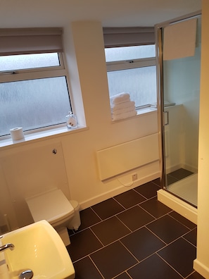 Baño con inodoro, lavabo y ducha. Cuenta con ventanas con persianas, un perchero y un pequeño estante con toallas.