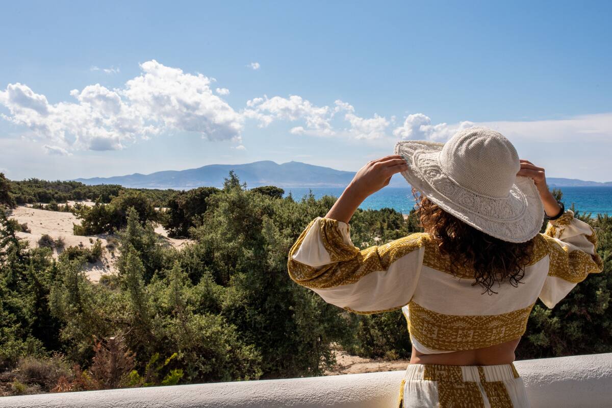 Une personne portant un chapeau blanc et un haut orné de motifs, qui surplombe un paysage côtier avec des arbres et des montagnes à l’horizon.
