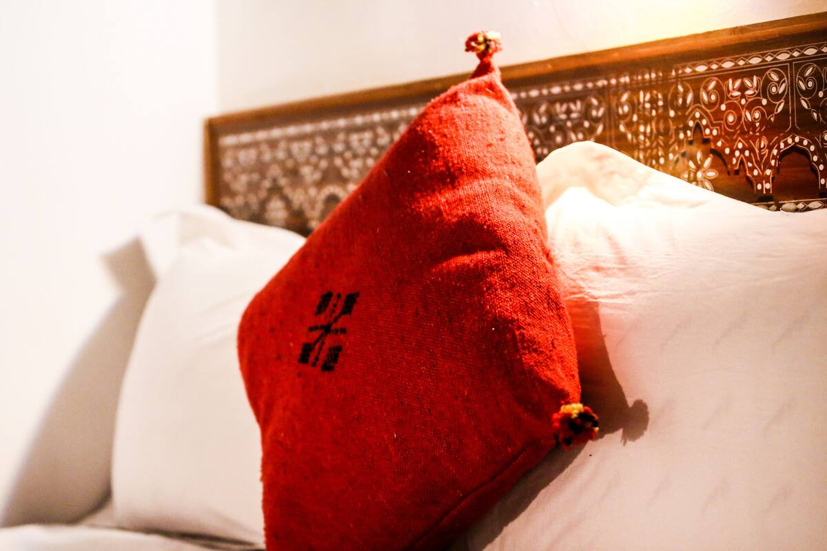 Una cama con un almohadón rojo y uno blanco.