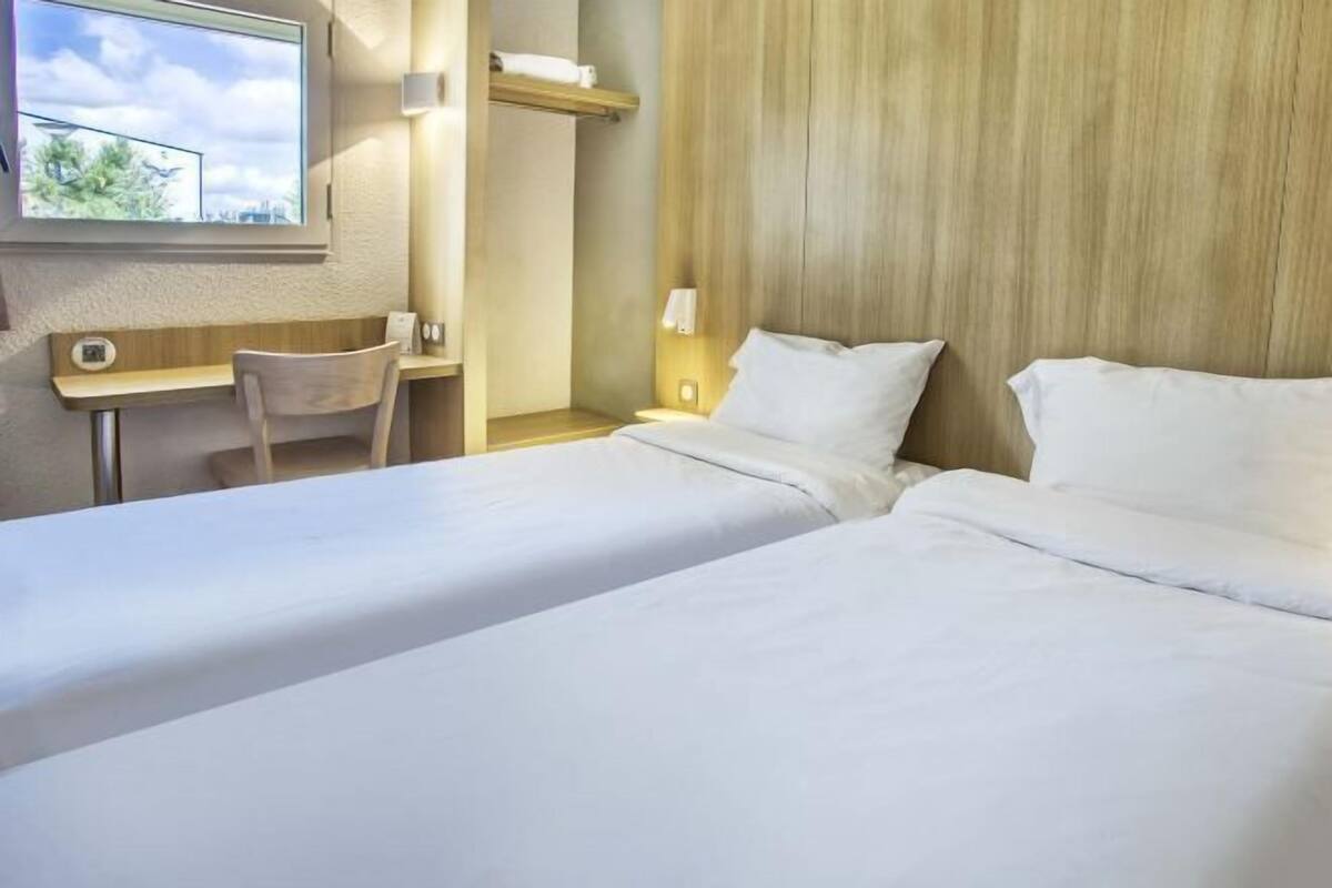 A hotel room with two beds, a desk, a chair, a window, and a wall-mounted shelf.