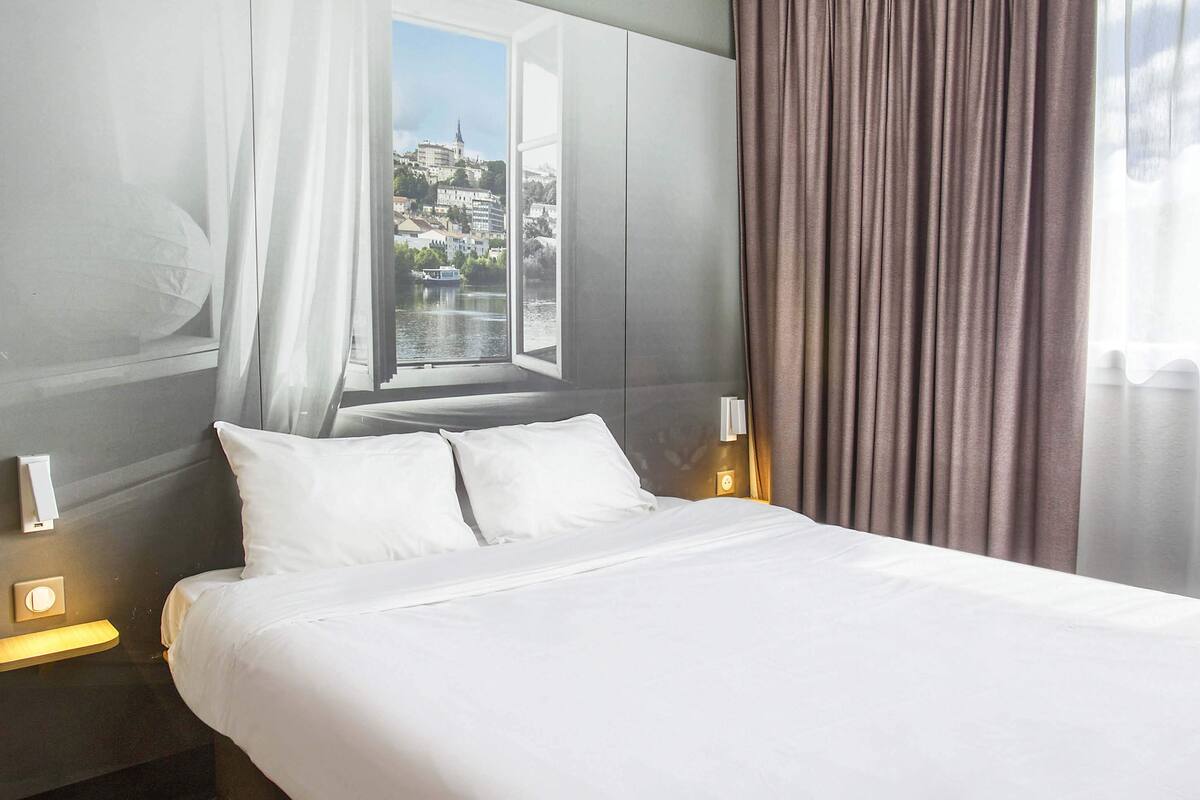 A hotel room with a large bed, a window with a view of a cityscape, and a curtain.
