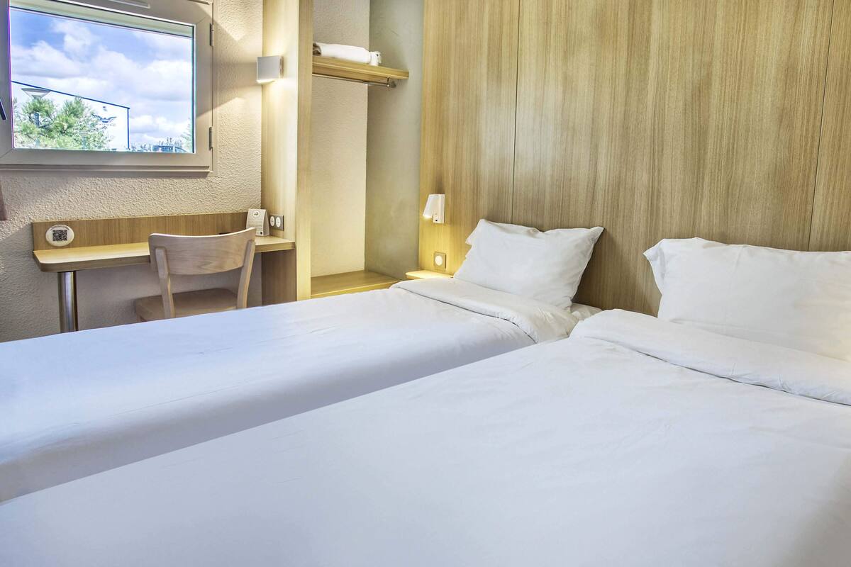 A hotel room with two beds, a desk, a chair, and a window with a view of the sky and trees.