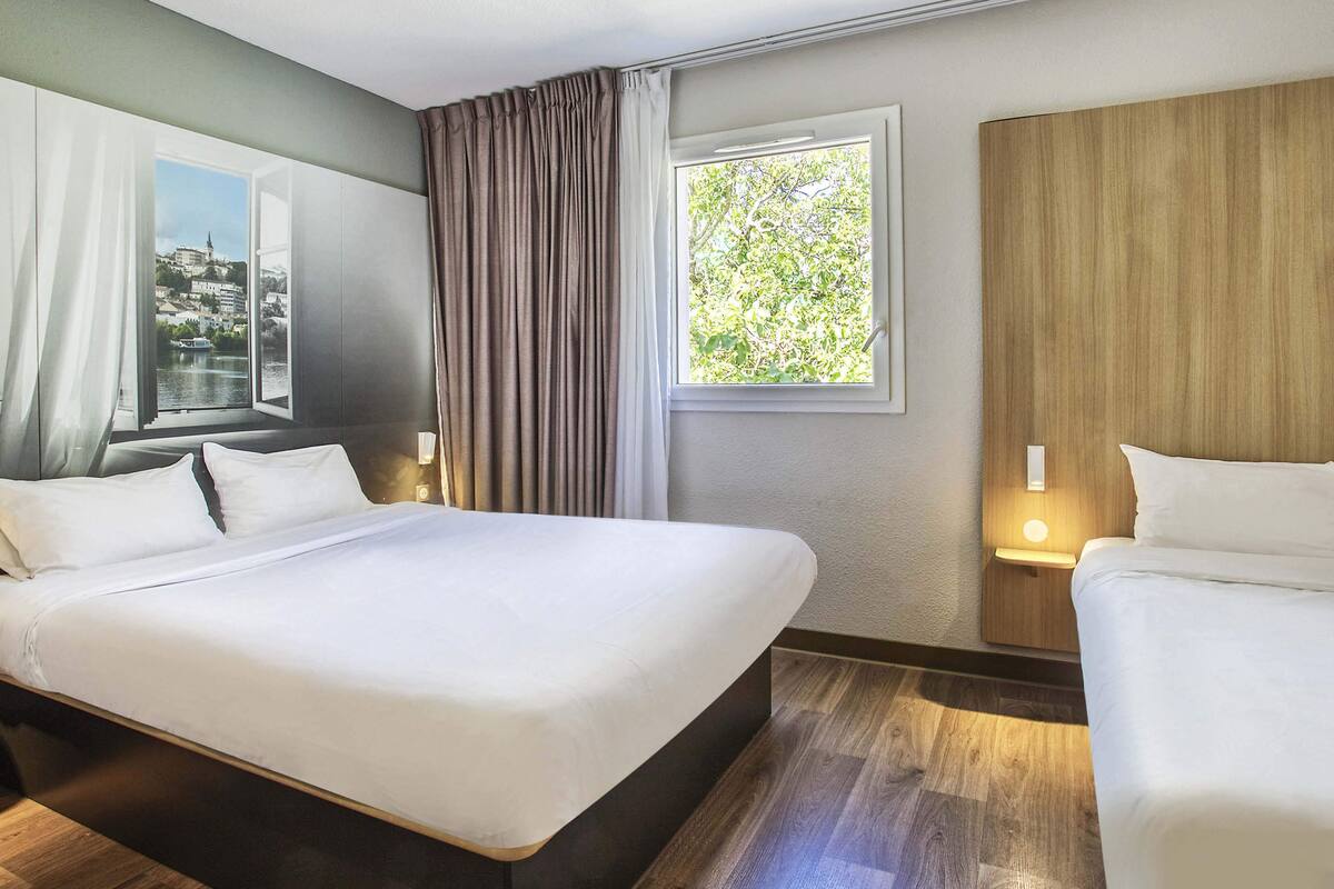 A hotel room with a large bed, a smaller bed, a window with a view of greenery, and a nightstand with a lamp.