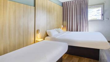 A hotel room with two single beds, wooden headboards, a window with curtains, and a small bedside table with a lamp.