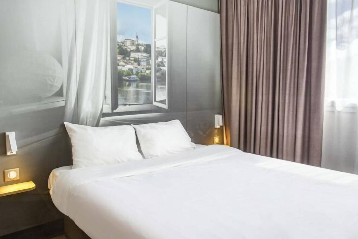 A hotel room with a bed, a window with curtains, a view of a cityscape, and a nightstand with a lamp.