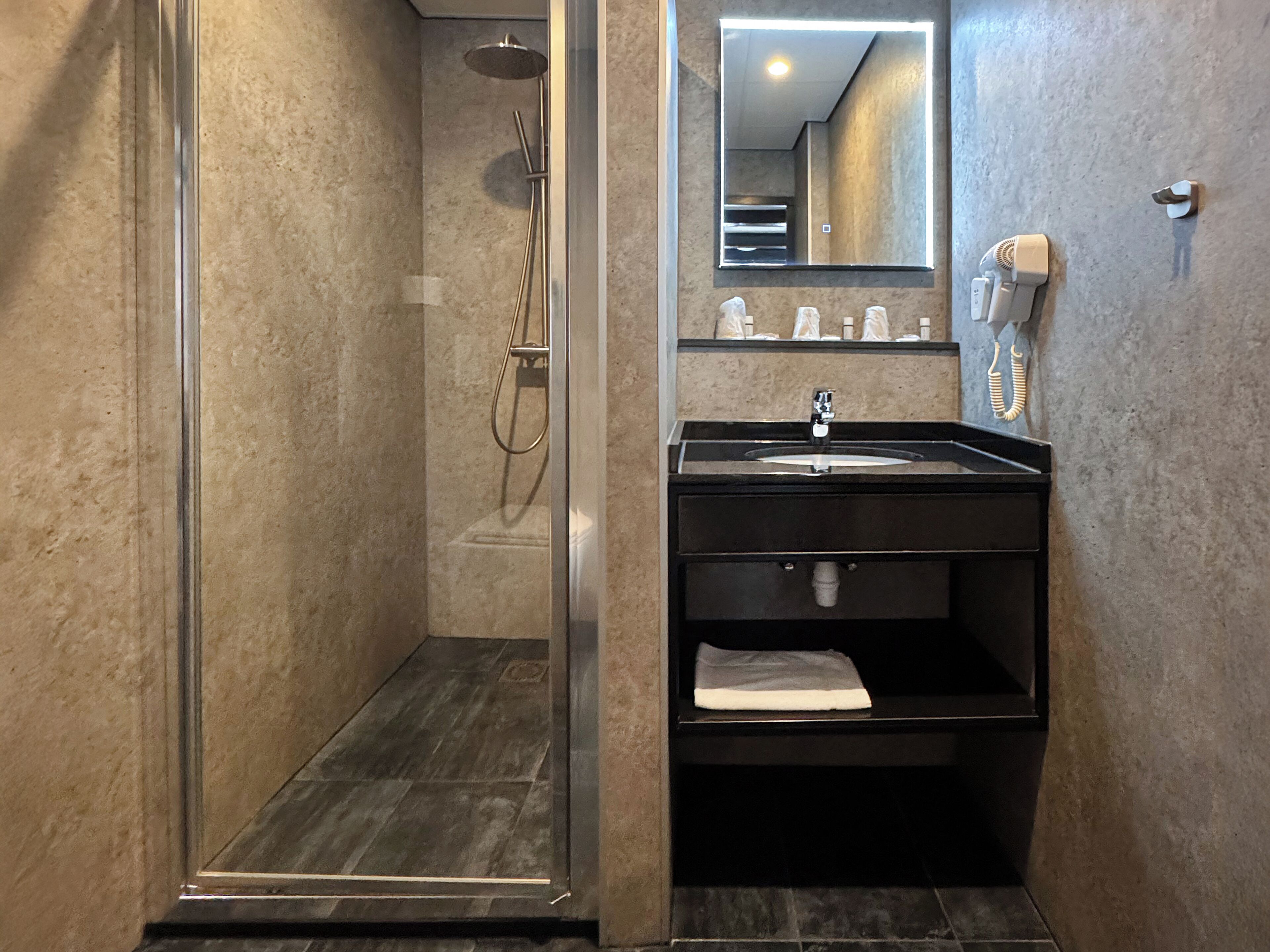 Un baño con ducha acristalada, un lavabo negro con un espejo encima y un secador de pelo en la pared.