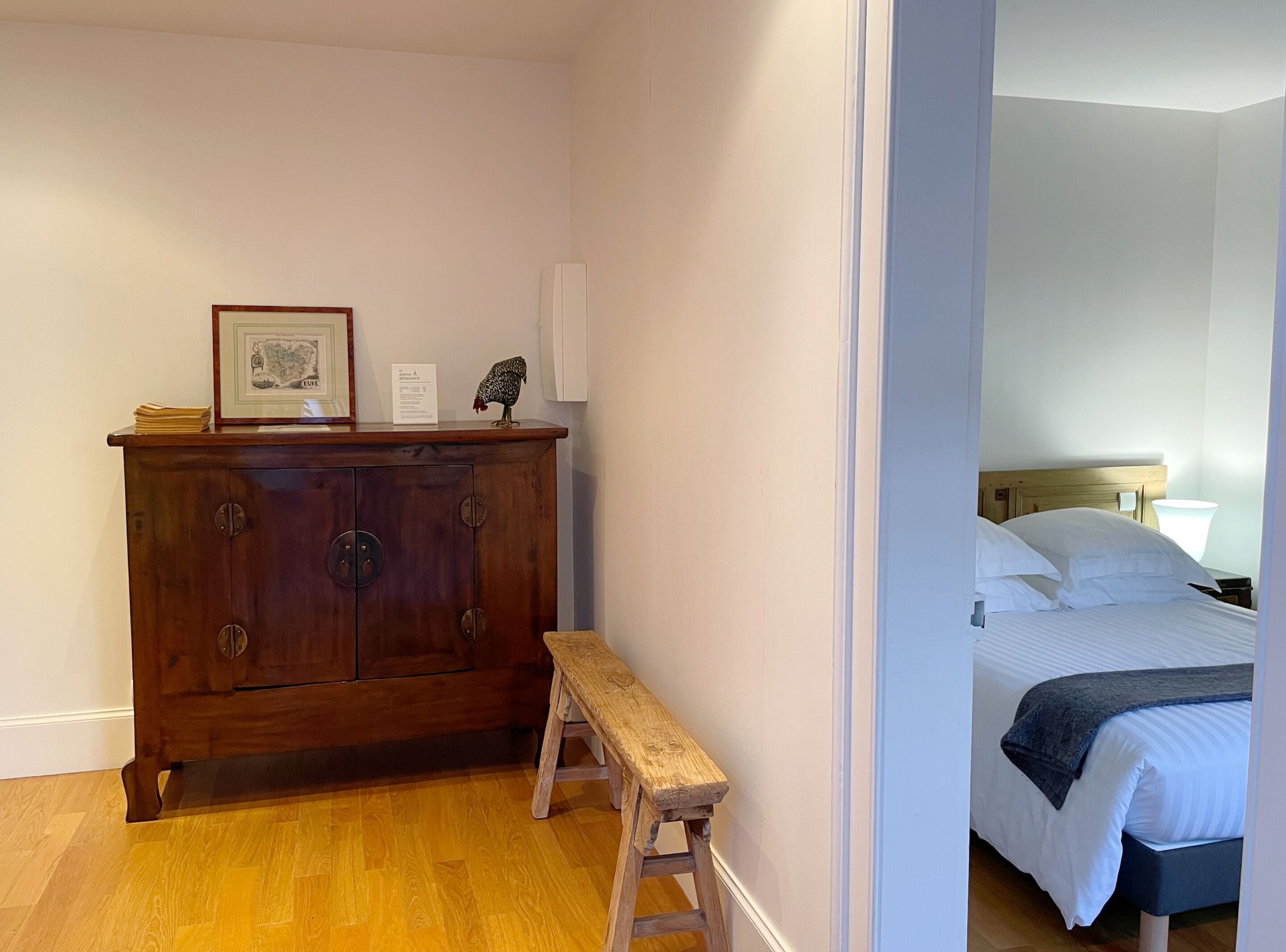 Una habitación con un aparador de madera, un mapa enmarcado, una figurita de gallo y un taburete de madera.