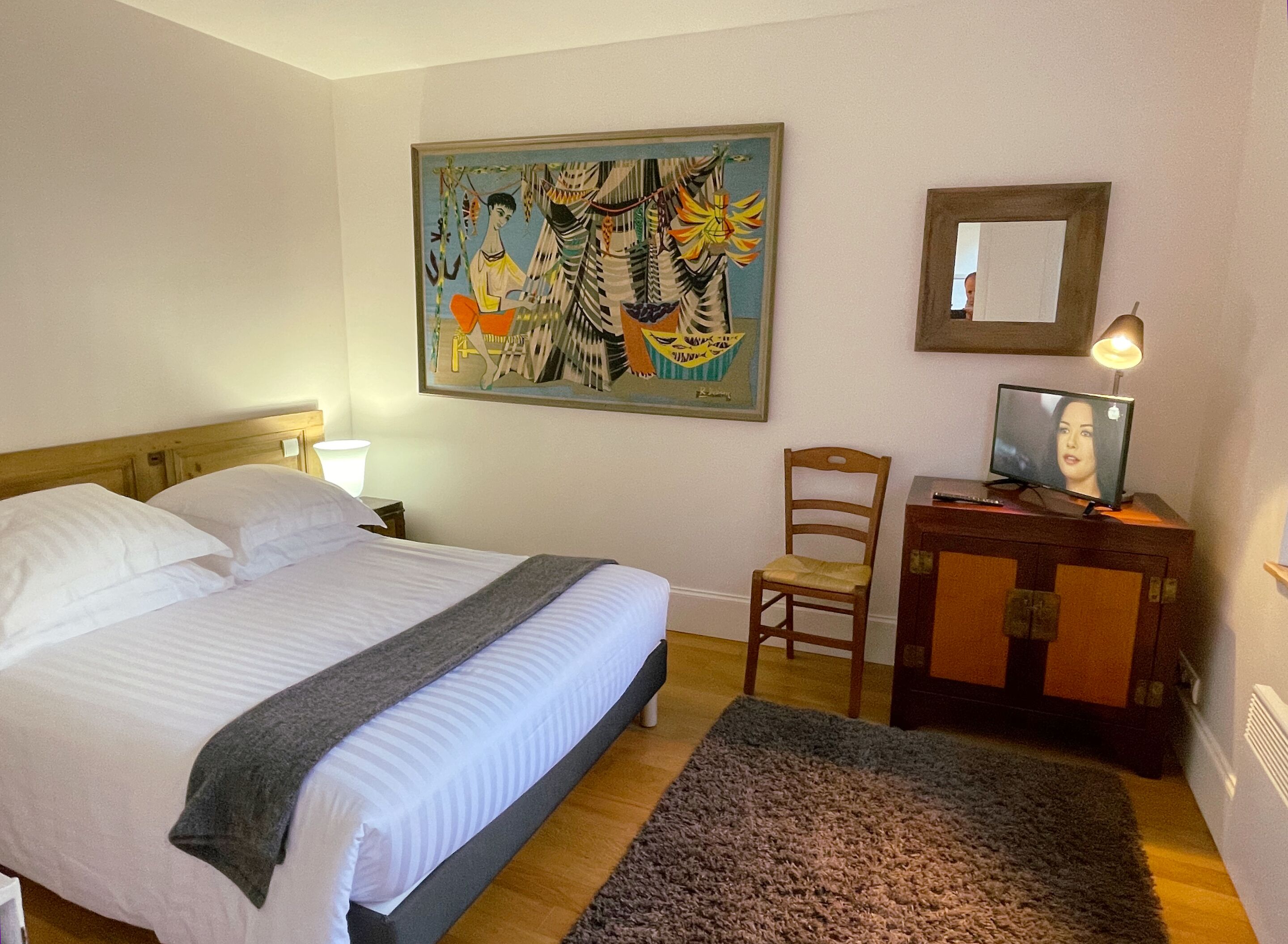 Ett hotellrum med en säng, ett skrivbord med en dator, en stol, en tavla på väggen och en lampa.