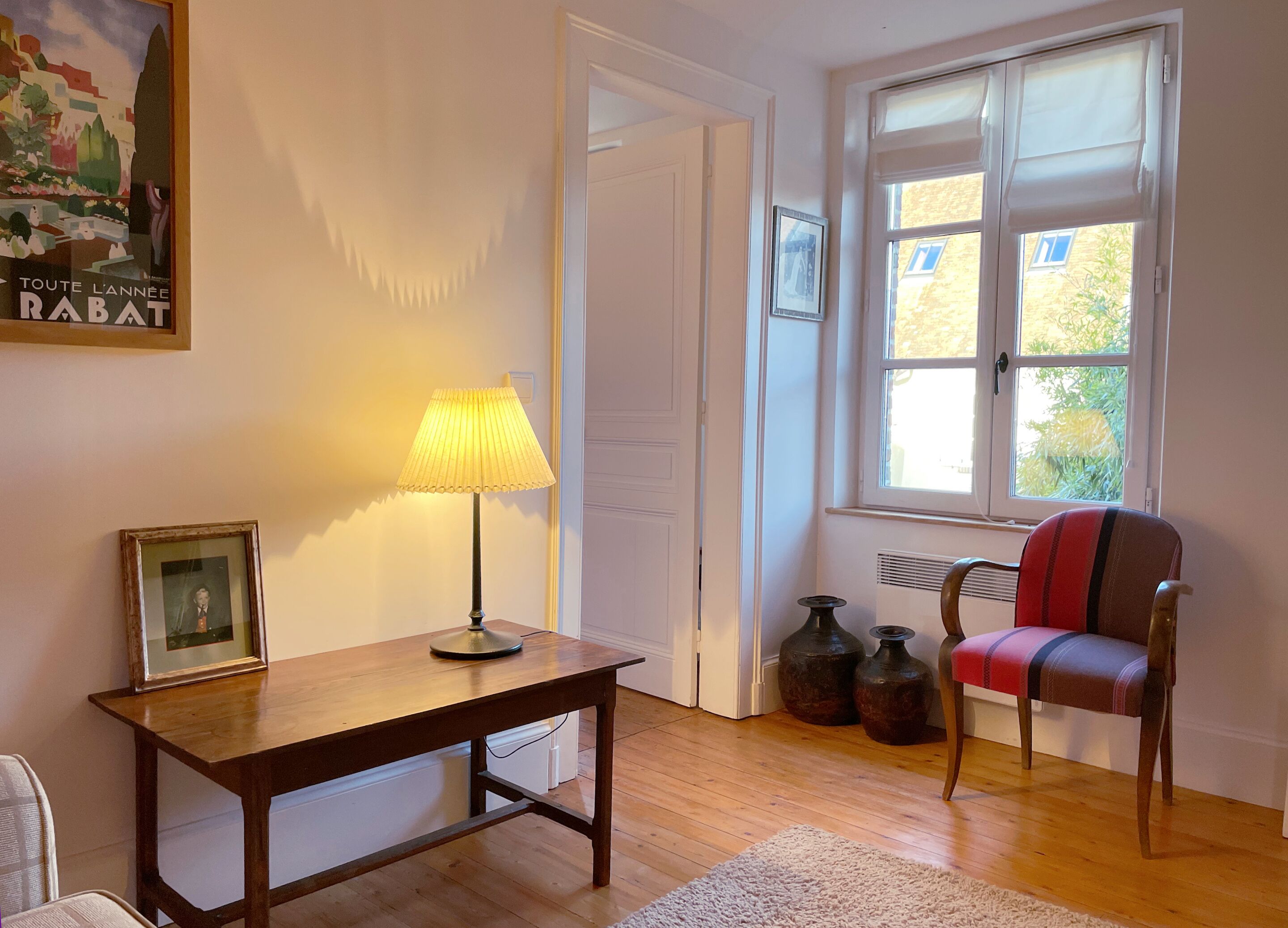 A room with a wooden table, a lamp, a chair, a radiator, and a window with a view of greenery.