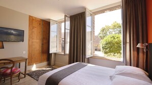 A hotel room with a bed, a desk, a chair, a window with curtains, and a painting on the wall. - Le Jardin Découverte (Conches-en-Ouche)