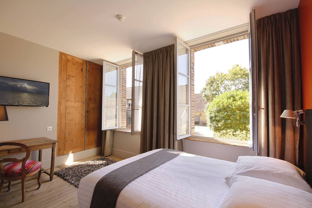 A hotel room with a bed, a desk, a chair, a window with curtains, and a painting on the wall.