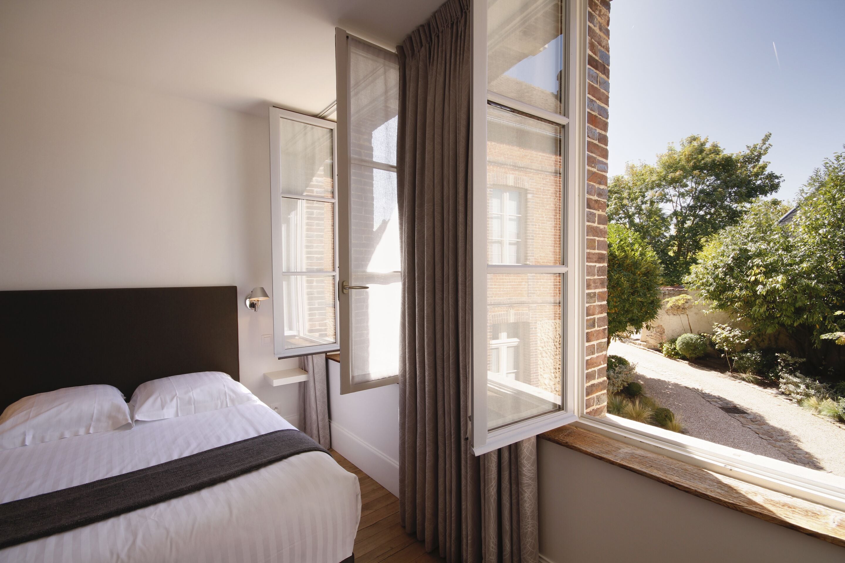 A bedroom with a large bed, a window with curtains, and a view of a garden and street.