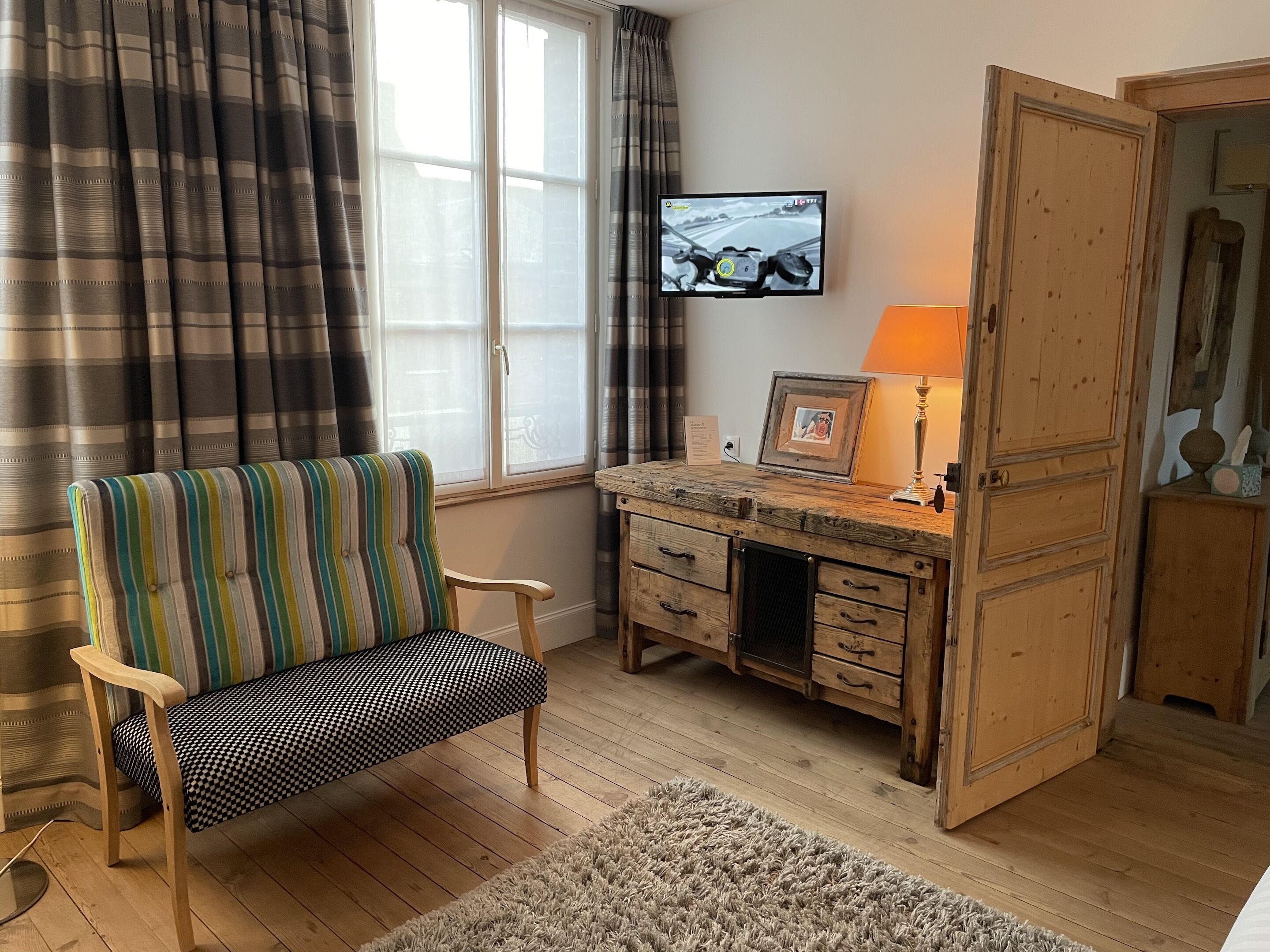 A cozy room with a striped chair, a wooden desk with drawers, a television mounted on the wall, and a large wooden door.