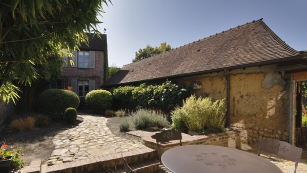 A courtyard with a stone pathway leading to a table and chairs, surrounded by greenery and a rustic building with a tiled roof. - Le Jardin Découverte (Conches-en-Ouche)