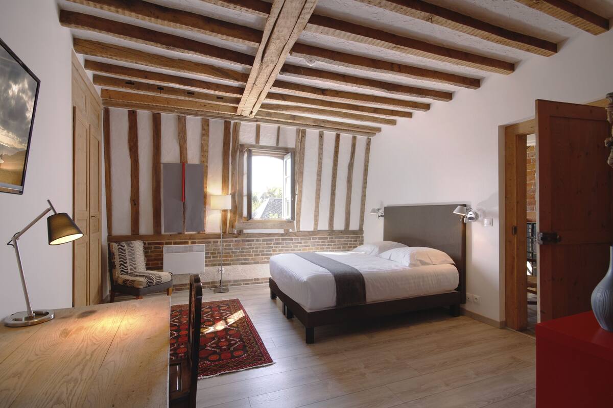 A bedroom with a wooden ceiling, a bed, a desk, a chair, a lamp, and a window.