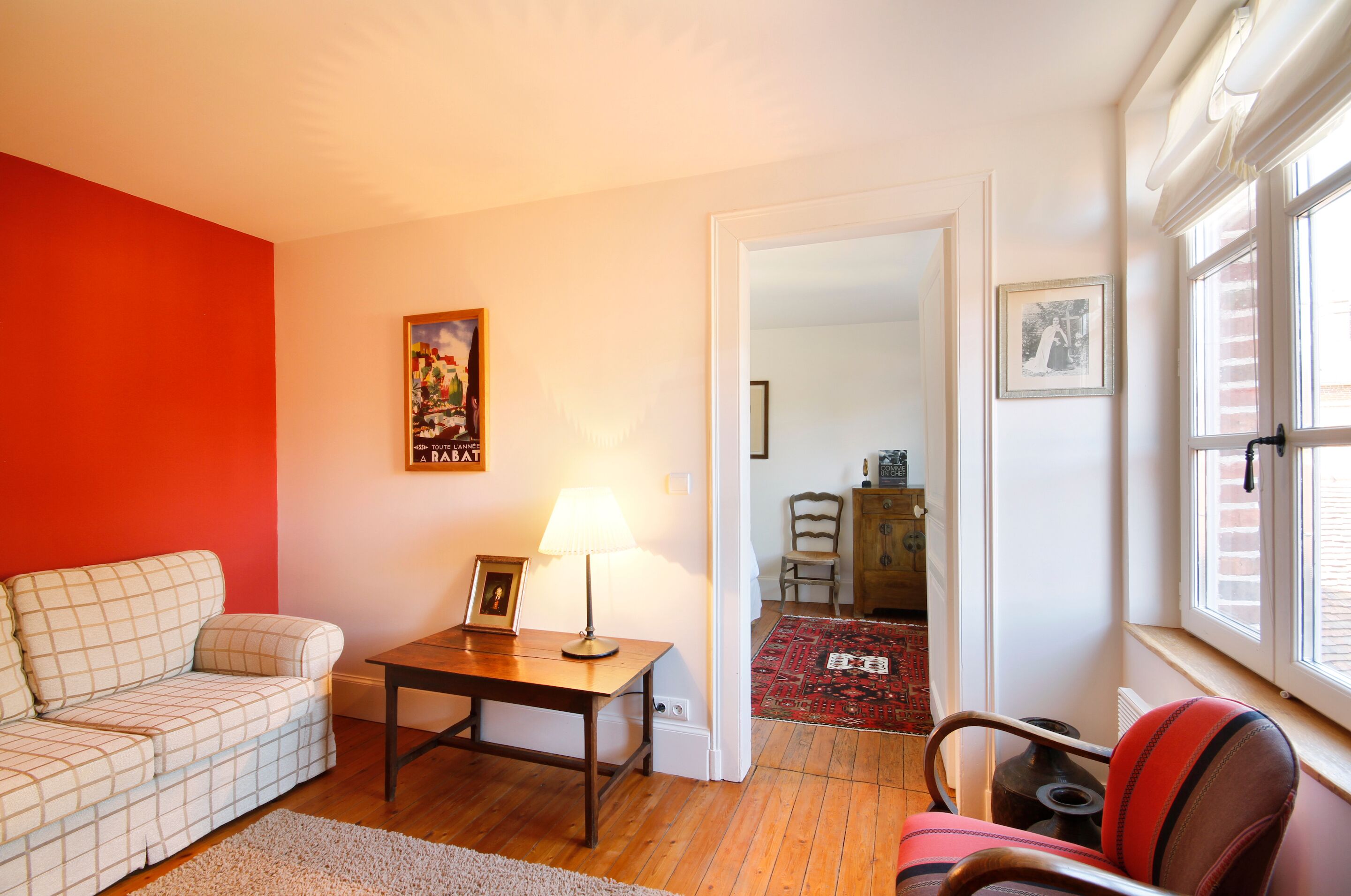 A living room with a red wall, a sofa, a table with a lamp, and a chair. There is a framed picture on the wall and a window with blinds.