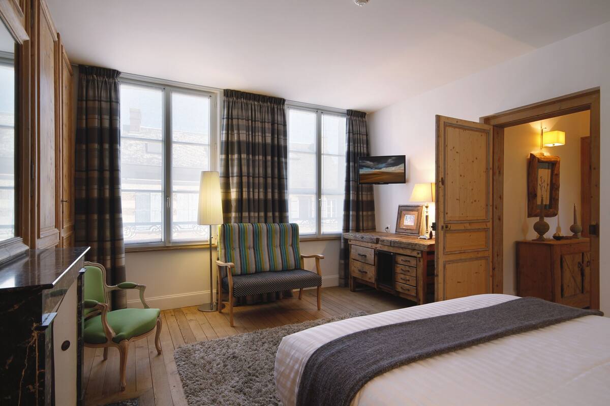 A hotel room with a bed, a desk with a chair, a window with curtains, and a wooden wardrobe.
