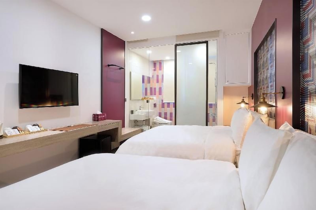 A hotel room with a bed, a TV, a desk, a mirror, and a bathroom visible through a glass door.