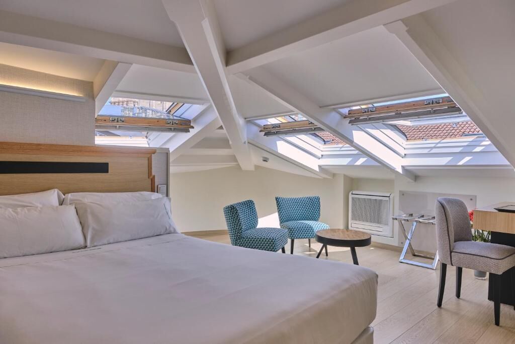 Una habitación de hotel moderna con una cama grande, ventanas tipo tragaluz, un área de estar con sillas y una pequeña mesa.