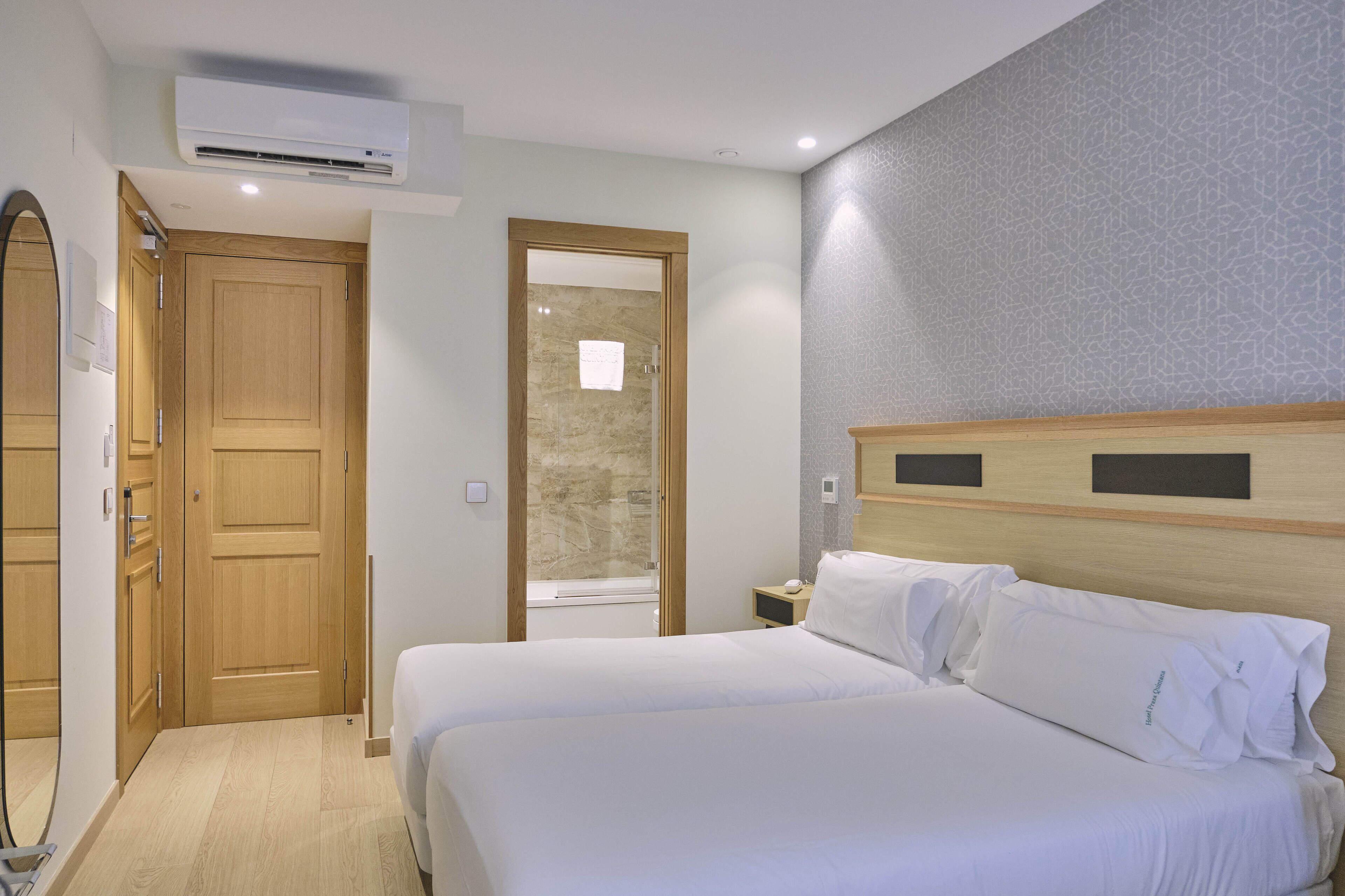 Habitación de hotel con una cama grande, un armario de madera, un baño con puerta de vidrio y un papel tapiz estampado.