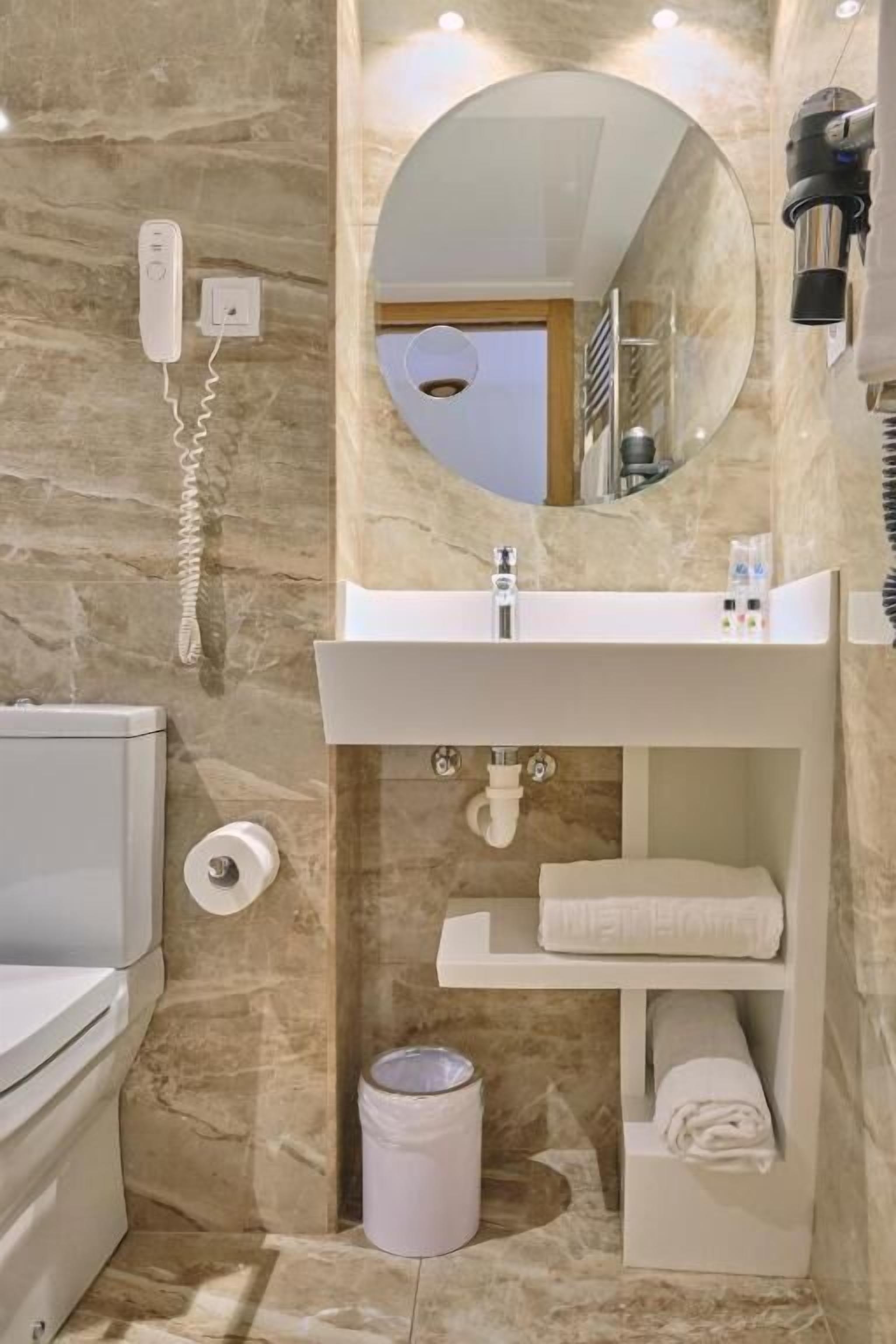 A bathroom with a marble wall, a white sink, a mirror, a towel rack, and a trash bin.