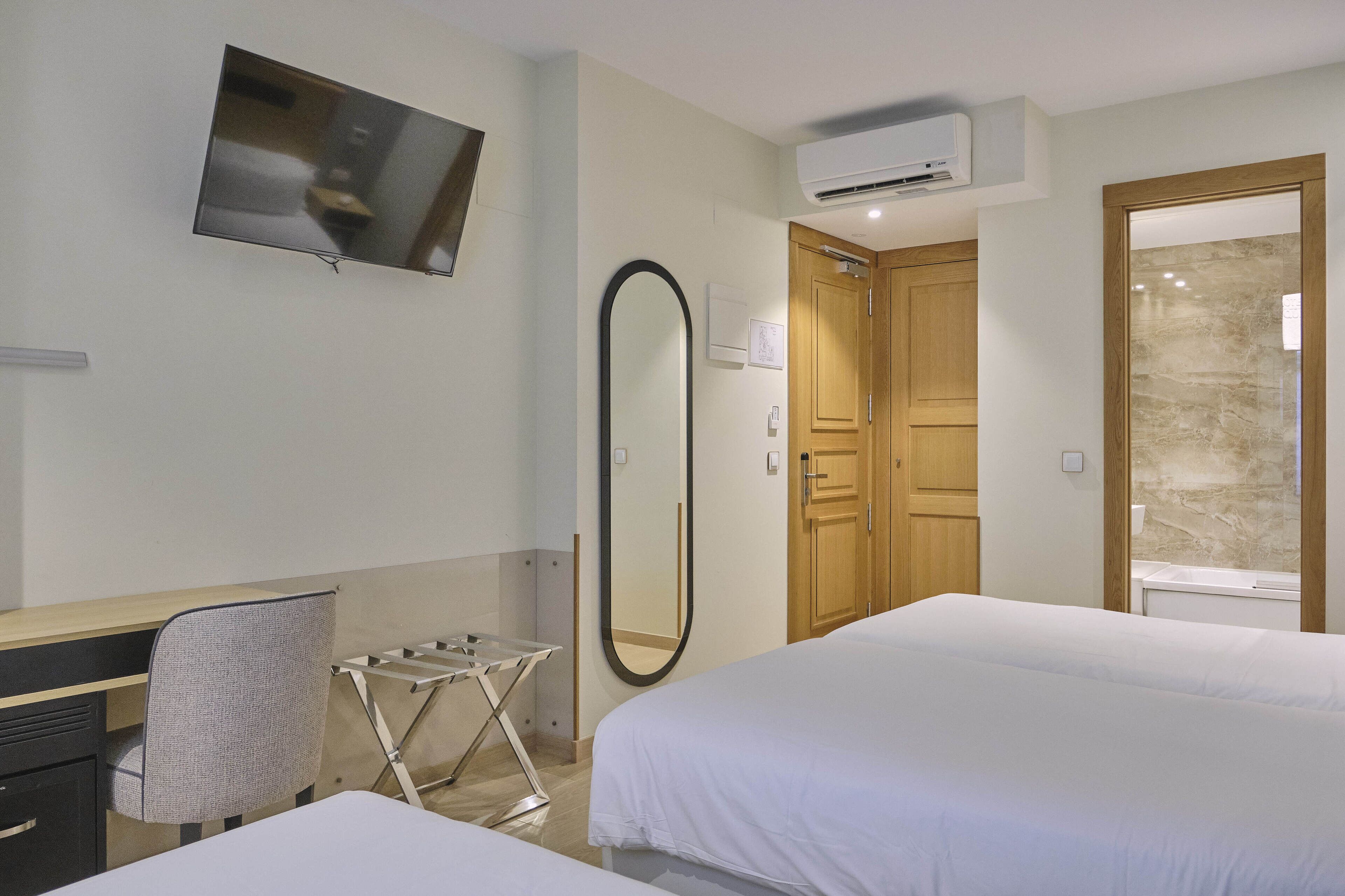 A hotel room with a bed, a desk with a chair, a TV mounted on the wall, a mirror, and a wooden door.