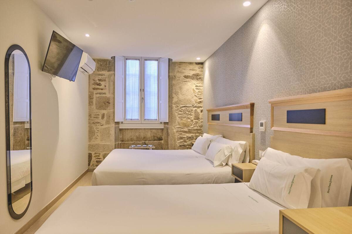 A hotel room with two beds, a stone wall, a TV mounted on the wall, and a window with curtains.
