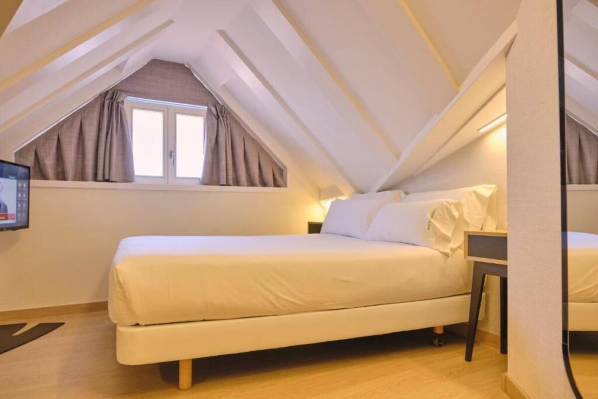A bedroom with a bed, a nightstand, a window with curtains, and a TV mounted on the wall.