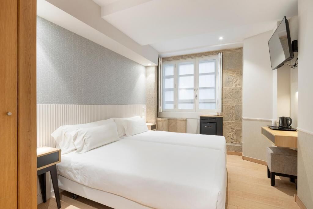 A modern hotel room with a large bed, a desk, a chair, a TV, and a window with a stone wall.