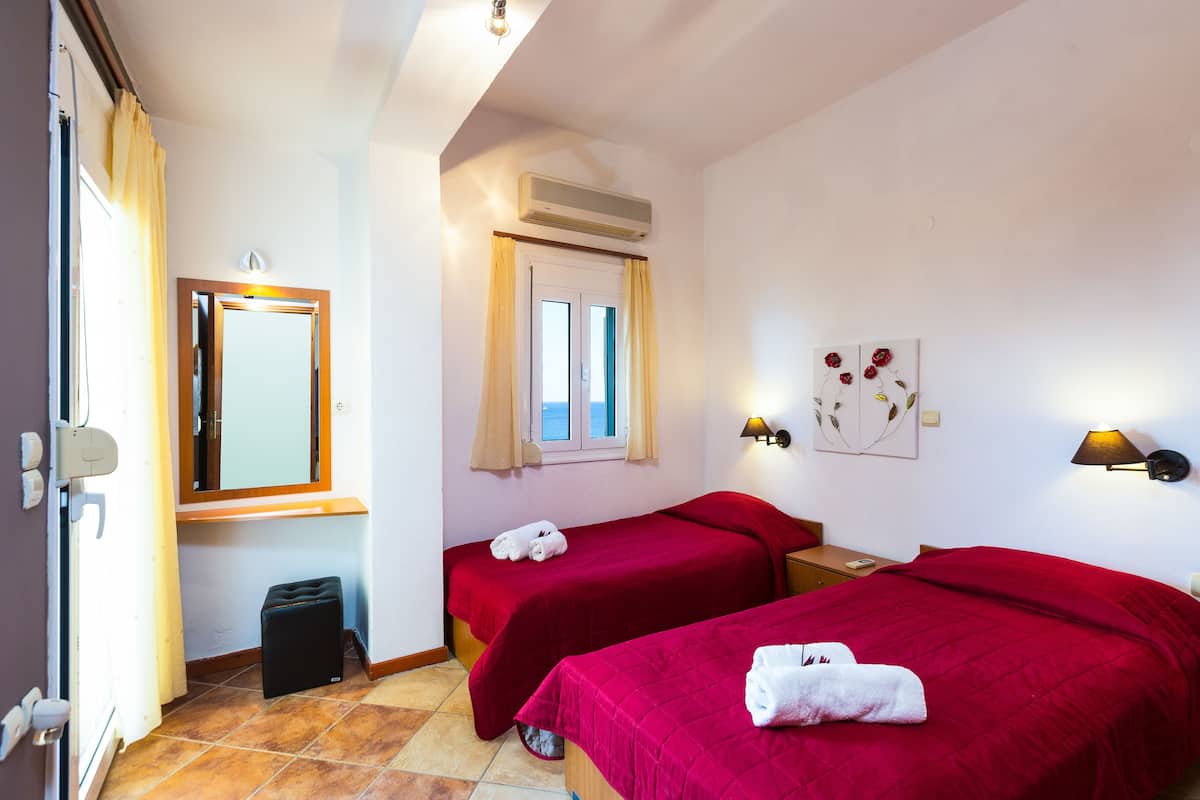 A hotel room with two beds, a window with curtains, an air conditioning unit, a wall-mounted lamp, and a small trash bin.