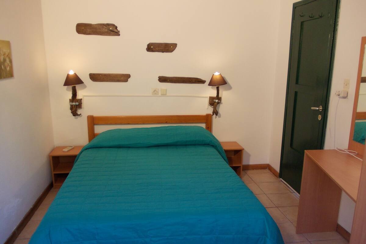 A bedroom with a wooden bed, teal bedding, two bedside lamps, a wooden nightstand, a green door, and a small wooden shelf.