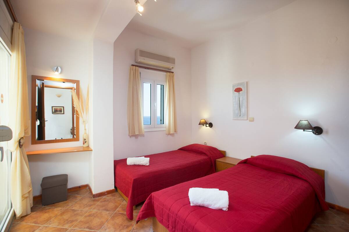 A hotel room with two beds, a window with curtains, a wall-mounted air conditioner, a mirror, and a small bedside table with a lamp.