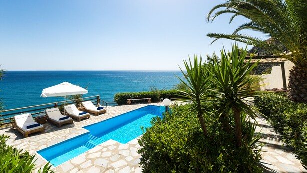 A pool with lounge chairs and umbrellas, surrounded by greenery and overlooking the sea. - Preveli Villas (Agios Vasileios)