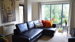 A modern living room with a black leather sectional sofa, a large window with a view of greenery, and a small round table with a plant. - MetroStays - Riverside Apartment (Dublin)