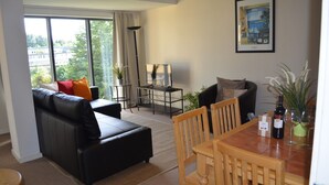 A living room with a black leather sofa, wooden dining table, and chairs, a TV on a stand, and a large window with curtains. - MetroStays - Riverside Apartment (Dublin)