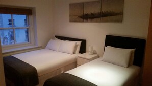 A hotel room with two single beds, a nightstand with a lamp, a window with a view of buildings, and a cityscape painting on the wall. - MetroStays - Stephen's Green 7-3 (Dublin)