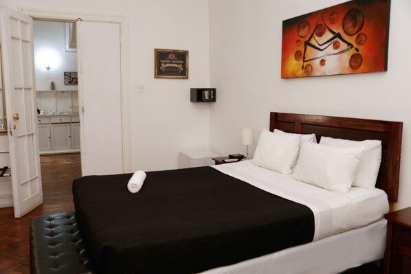 Habitación de hotel con cama, mesita de noche, lámpara de noche, un cuadro en la pared y vistas a la cocina.