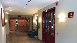 Et hotellets receptionområde med en receptionsdisk, et siddeområde med en stol og en potteplante, og en rød dør med glaspaneler.