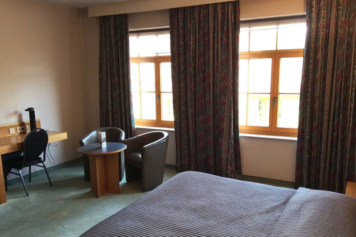 A hotel room with a bed, a desk with a chair, a small table with a chair, and a window with curtains.