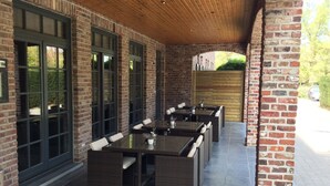 En udendørs patio med murstensvægge og store vinduer, med flere borde og stole.
