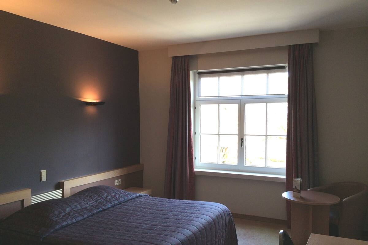 A hotel room with a bed, a small table, a chair, a window with curtains, and a wall-mounted light.