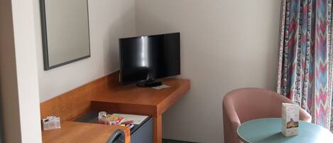 A hotel room with a wooden desk, a television, a small table with a cup, a chair, and a bed.