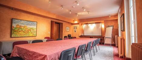 Un salón de conferencias con una larga mesa, sillas, una pantalla de proyector y un cuadro en la pared.