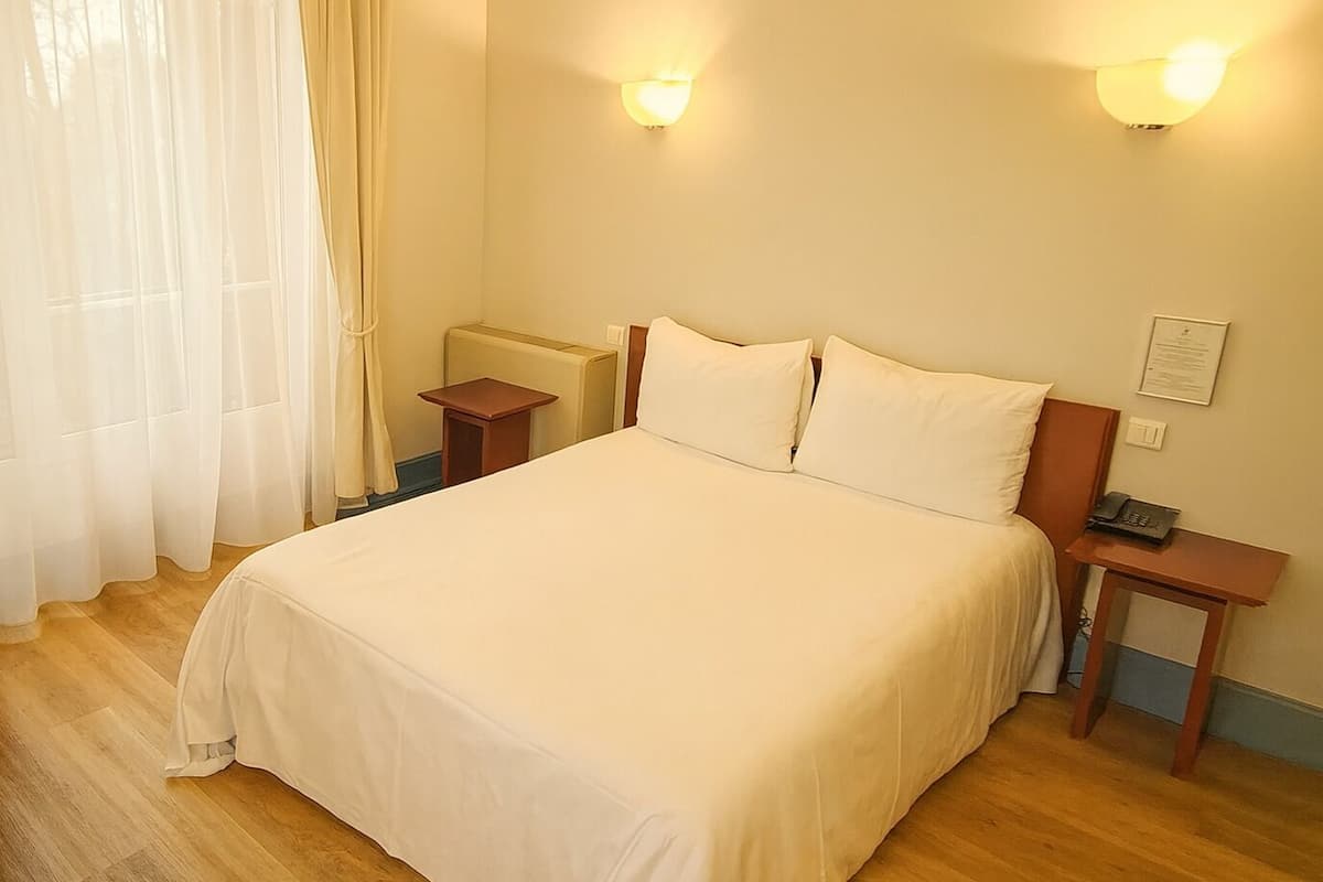 A neatly made bed in a hotel room with a wooden headboard, two bedside tables, and a wall-mounted light fixture.